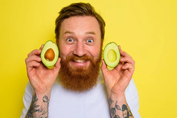 Uomo felice con la barba e tatuaggi tiene un avocado — Foto Stock