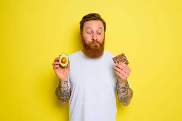 Douter homme à la barbe et tatouages tient avocat et chocolat — Photo