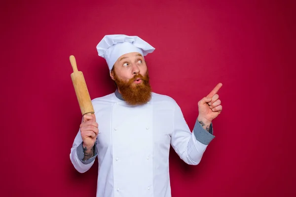 Chef stupito con barba e grembiule rosso chef tiene mattarello in legno — Foto Stock