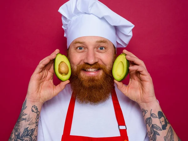 Szczęśliwy szef kuchni z brodą i czerwony fartuch posiada awokado — Zdjęcie stockowe
