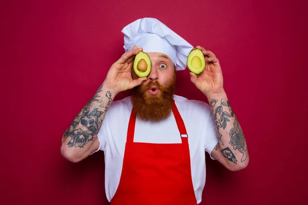 Surprised chef with beard and red apron holds an avocado — Zdjęcie stockowe