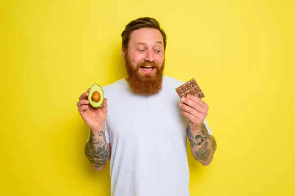 Lycklig man med skägg och tatueringar rymmer avokado och choklad — Stockfoto