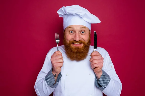 Szczęśliwy szef kuchni z brodą i czerwony fartuch trzyma sztućce w dłoni — Zdjęcie stockowe