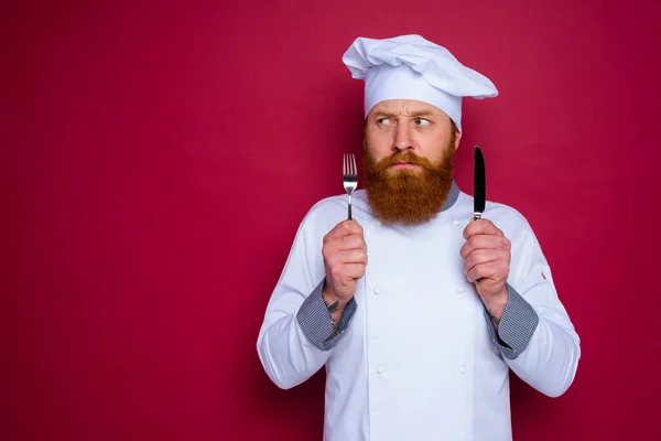 Pensoso chef con barba e grembiule rosso tiene in mano le posate — Foto Stock