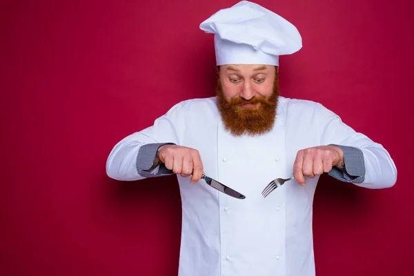 Wątpiący szef kuchni z brodą i czerwonym fartuchem trzyma sztućce w dłoni — Zdjęcie stockowe