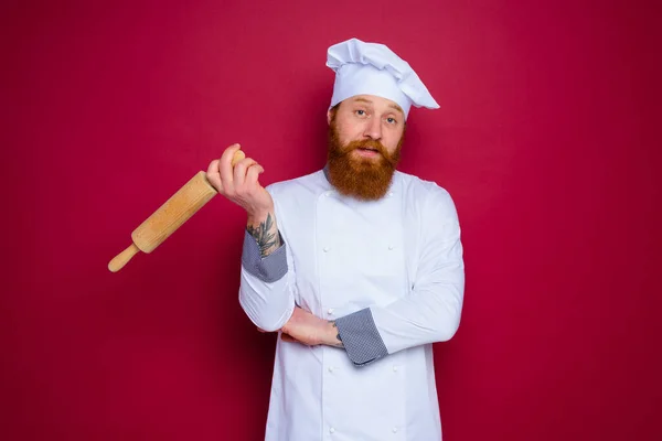 Cuoco pensoso con barba e grembiule rosso chef tiene mattarello in legno — Foto Stock