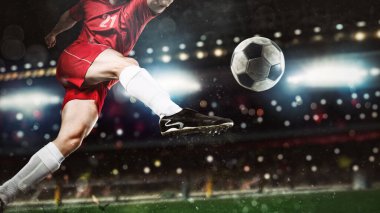 Gece bir futbol maçının kapanış sahnesinde kırmızı üniformalı bir oyuncu topa güçlü bir şekilde vuruyor.