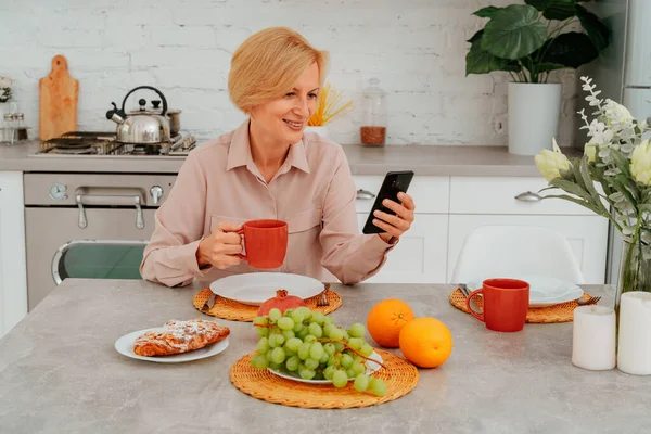 Vrouw ontbijt thuis met fruit, cake en koffie en leest nieuws van smartphone — Stockfoto