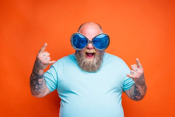 Tlustý šťastný muž s vousy, tetováním a slunečními brýlemi dělá gesto rohů — Stock fotografie