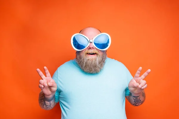 Grasso uomo felice con la barba, tatuaggi e occhiali da sole fa il segno della vittoria con le mani — Foto Stock