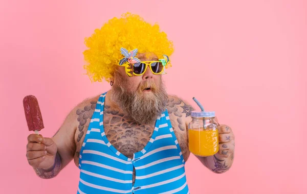 Grasso pensoso uomo con barba e parrucca mangia un ghiacciolo e beve un succo di frutta — Foto Stock