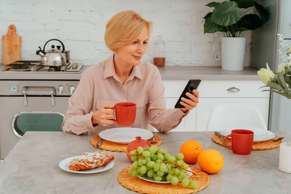 Vrouw ontbijt thuis met fruit, cake en koffie en leest nieuws van smartphone — Stockfoto