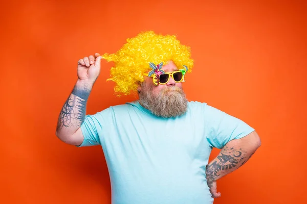 Tłusty szczęśliwy mężczyzna z brodą, tatuażami i okularami przeciwsłonecznymi zabawia się żółtą peruką — Zdjęcie stockowe