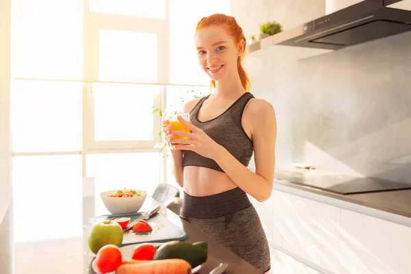 Молодая спортсменка с рыжими волосами на кухне со стаканом фруктового центрифужного сока — стоковое фото