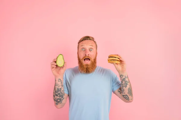 Niesamowity człowiek z brodą i tatuażami jest niezdecydowany czy zjeść awokado czy hamburgera — Zdjęcie stockowe