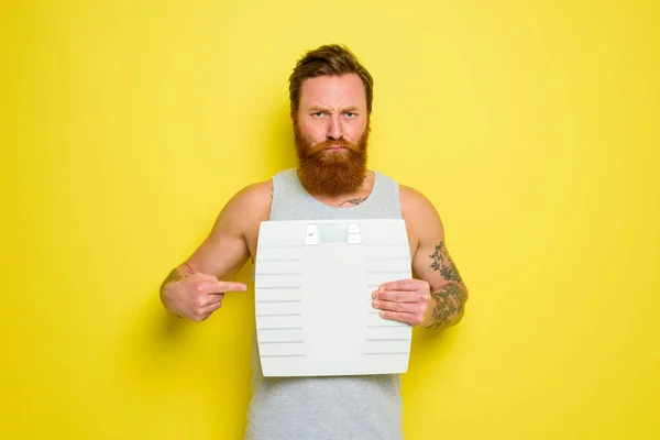 Unhappy man with beard and tattoos holds an electronic balance — Stock Photo, Image
