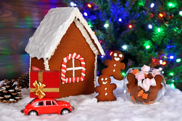 手作りの食べられるジンジャーブレッドハウス 小さな男性とお菓子 雪の装飾 ギフト付きの車 背景にガーランド付きの新年の木 — ストック写真