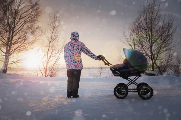 Mãe com um bebê em um carrinho de bebê no inverno ao pôr do sol em um dia gelado — Fotografia de Stock