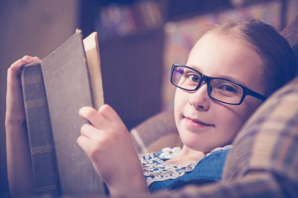 Flicka som läser en bok hemma sitter i en fåtölj. — Stockfoto