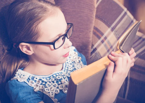 Flicka som läser en bok hemma sitter i en fåtölj. — Stockfoto