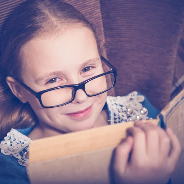 Flicka som läser en bok hemma sitter i en fåtölj. — Stockfoto