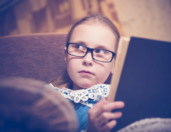 Flicka som läser en bok hemma sitter i en fåtölj. — Stockfoto