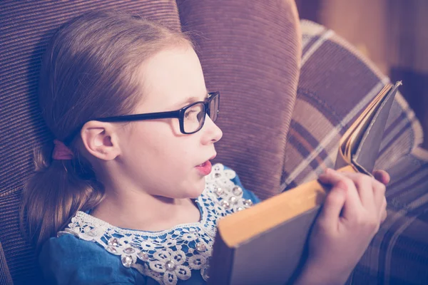 Flicka som läser en bok hemma sitter i en fåtölj. — Stockfoto