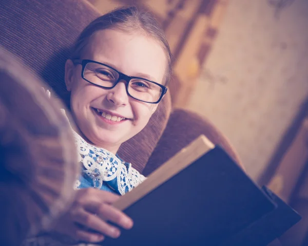 Flicka som läser en bok hemma sitter i en fåtölj. — Stockfoto