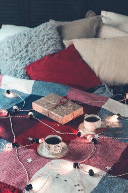 Kahve fincanları ve hediye kutuları renkli kareli battaniye ve hafif çelenk ve yıldızlarla süslenmiş çeşitli yastıklar ile rahat bir yatağa konur.