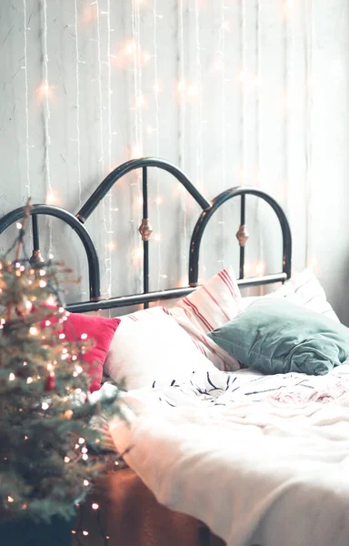 Retro styled unmade bed with metal headboard with light linens and green and red pillows in room with Christmas tree and luminous garland
