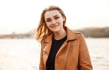 Havalı bir gecede gölün kıyısında dikilirken gülümseyen ve kameraya bakan şık deri ceketli mutlu genç bayan.