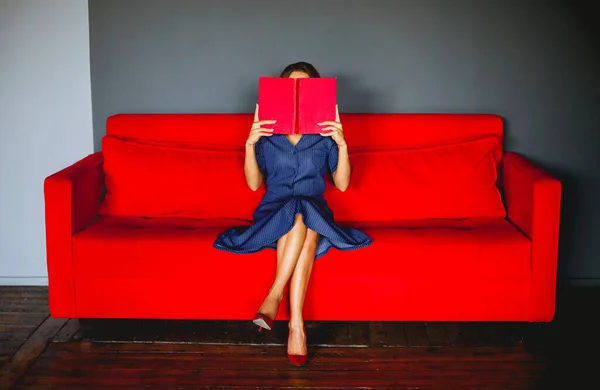 ソファに座って面白い本を楽しんでレトロなドレスを着て赤い唇を持つエレガントな女性 — ストック写真