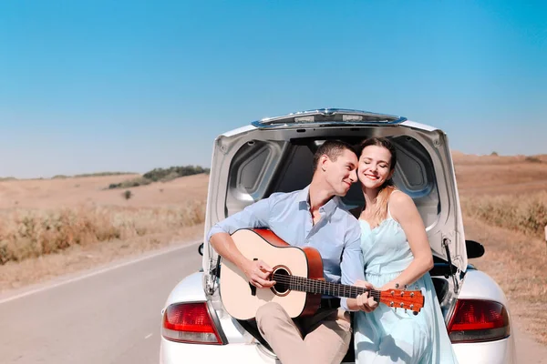 夏の旅行中に田舎道に停められた車のトランクに座っている間 幸せな若いカップルはギターを演奏し 一緒に時間を楽しむ — ストック写真