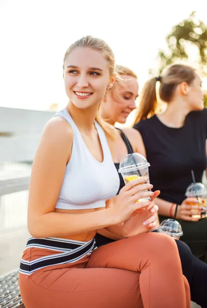 穿着运动服的年轻女性面带微笑 望着远方 一边享受着新鲜健康的饮料 一边坐在女友身边的长椅上 — 图库照片