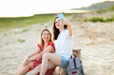 Neşeli bayan arkadaşlar kumlu sahilde oturur ve yazın akıllı telefonlarda selfie çekerler.