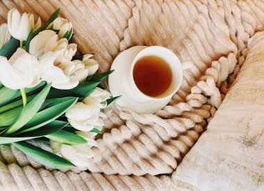 Taze beyaz lale buketi ve bej peluş battaniye ve yastıkla rahat bir yatağa yerleştirilmiş beyaz kahve fincanı.