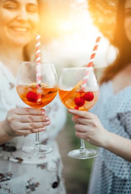 Kırpılmış kadın arkadaşlar, güneşli yaz gününde alkol bardaklarını kiraz ve pipetle tokuştururlar.