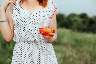 İsimsiz bir kadın, kırsal kesimin bulanık arka planında kirazlı ve çizgili saman şeritli alkol bardağı taşıyor.