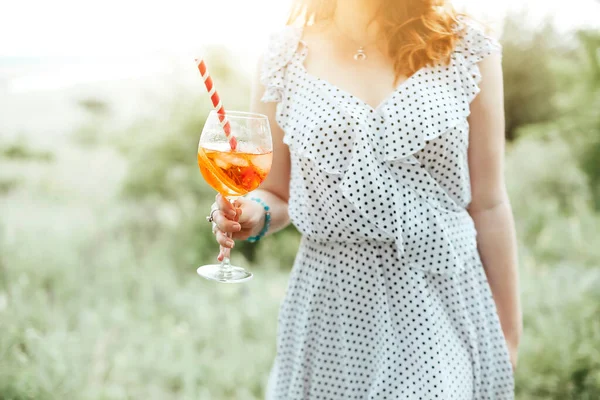 Young female drinking alcohol cocktail with orange on blurred background of nature