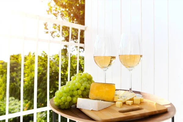 Glasses of wine placed on table near cutting board with ripe grapes and cheese on sunny day on terrace