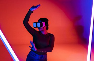 VR kulaklıklı mutlu kadın gelecekteki odada kırmızı neon ışığı altında gülümseyip sanal gerçekliği keşfediyor.