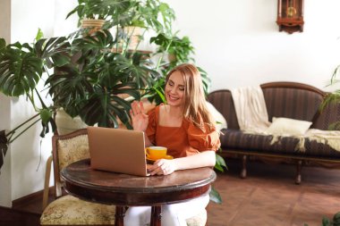 Neşeli kadın klasik masada oturuyor, oturma odasında dizüstü bilgisayarına el sallıyor.