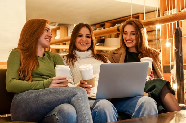 Cheerful women with coffee to go smiling and browsing laptop while resting in cafeteria in weekend together