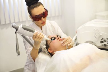 Ofisteki koltukta uzanan gözlüklü müşteriye lazer yüzeyi yapan odaklanmış kadın kozmetikçi.