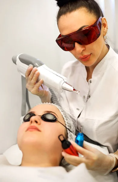 Focused female cosmetician doing laser resurfacing to client in glasses lying on couch in office