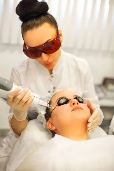 Focused female cosmetician doing laser resurfacing to client in glasses lying on couch in office