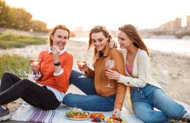Neşeli genç kız arkadaşlar, yaz tatilinde parkta piknik yaparken şarap içip bruschetta yiyorlar.
