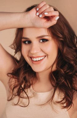 Moda kıyafetleri içinde, dalgalı saçlarıyla gülümseyen ve bej arka planda kameraya bakan neşeli genç bir kadın.