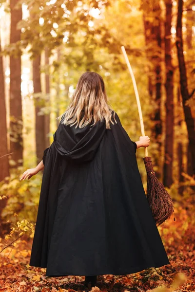 魔女の衣装を着た無名の女性のバックビューでは 森の中でハロウィーンの日に秋の葉で覆われたパスを歩いてほうき — ストック写真