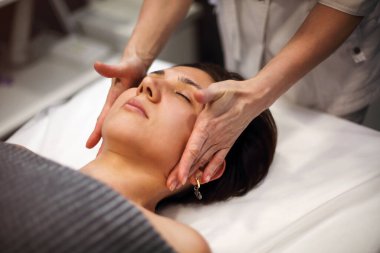 Gecelik giymiş, kaplıca salonunda duran ve kadın müşterinin yüzüne masaj yapan tanınmamış bir masöz.